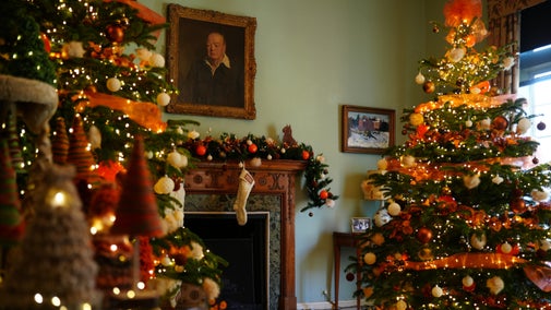 Yarn Christmas trees on a cat climbing tree, orange and white ribbons, baubles and balls of fluff decorate the Christmas Trees and garlands. A pair of white knitted stockings above the fireplace. A silhouette of a cat on the mantlepiece. Portrait of Winston Churchill on the wall and a winter landscape painting of Chartwell below.
